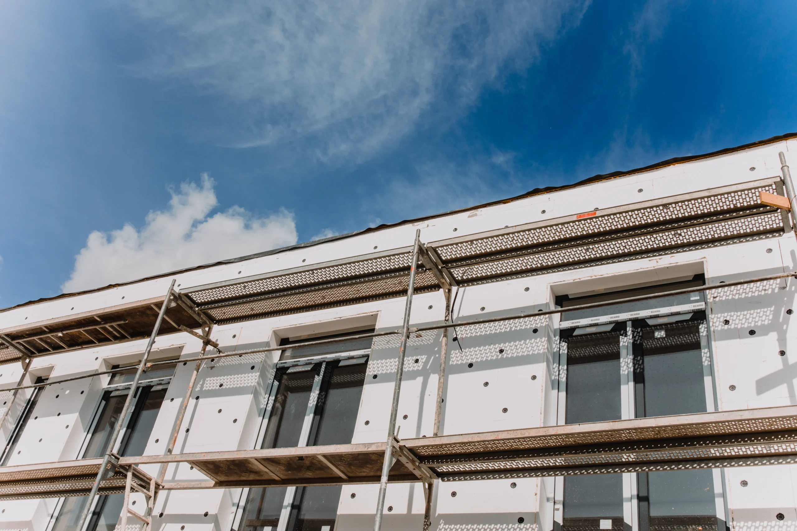 Façade d'un bâtiment en cours d'isolation thermique par l'extérieur (ITE). Pose de panneaux isolants blancs et échafaudages métalliques devant des fenêtres modernes.