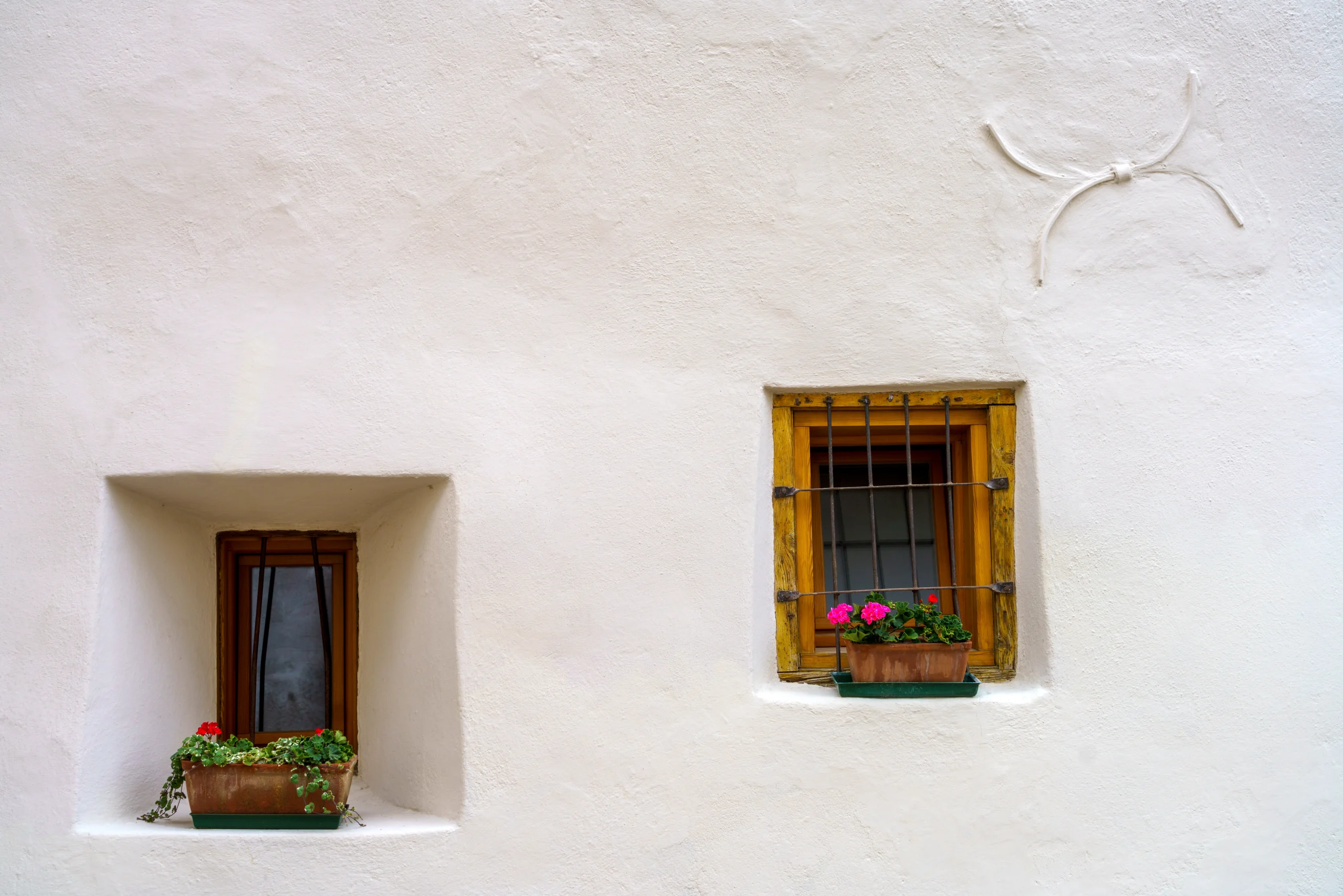 Deux petites fenêtres traditionnelles avec jardinières de géraniums rouges sur un mur blanc texturé.