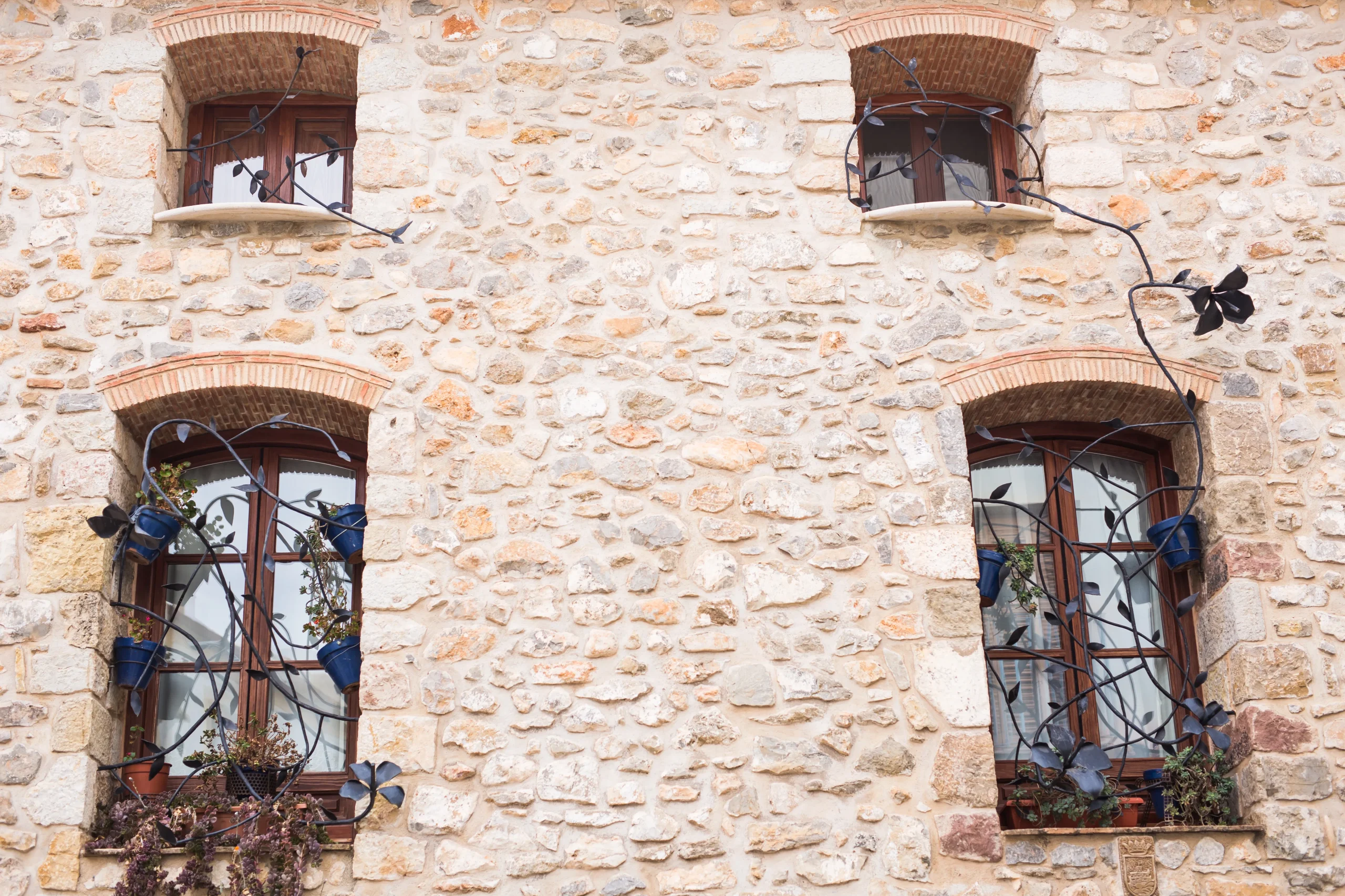 Façade en pierres apparentes jointoyées et briques anciennes autour de quatre fenêtres en bois avec grilles décoratives en fer forgé.