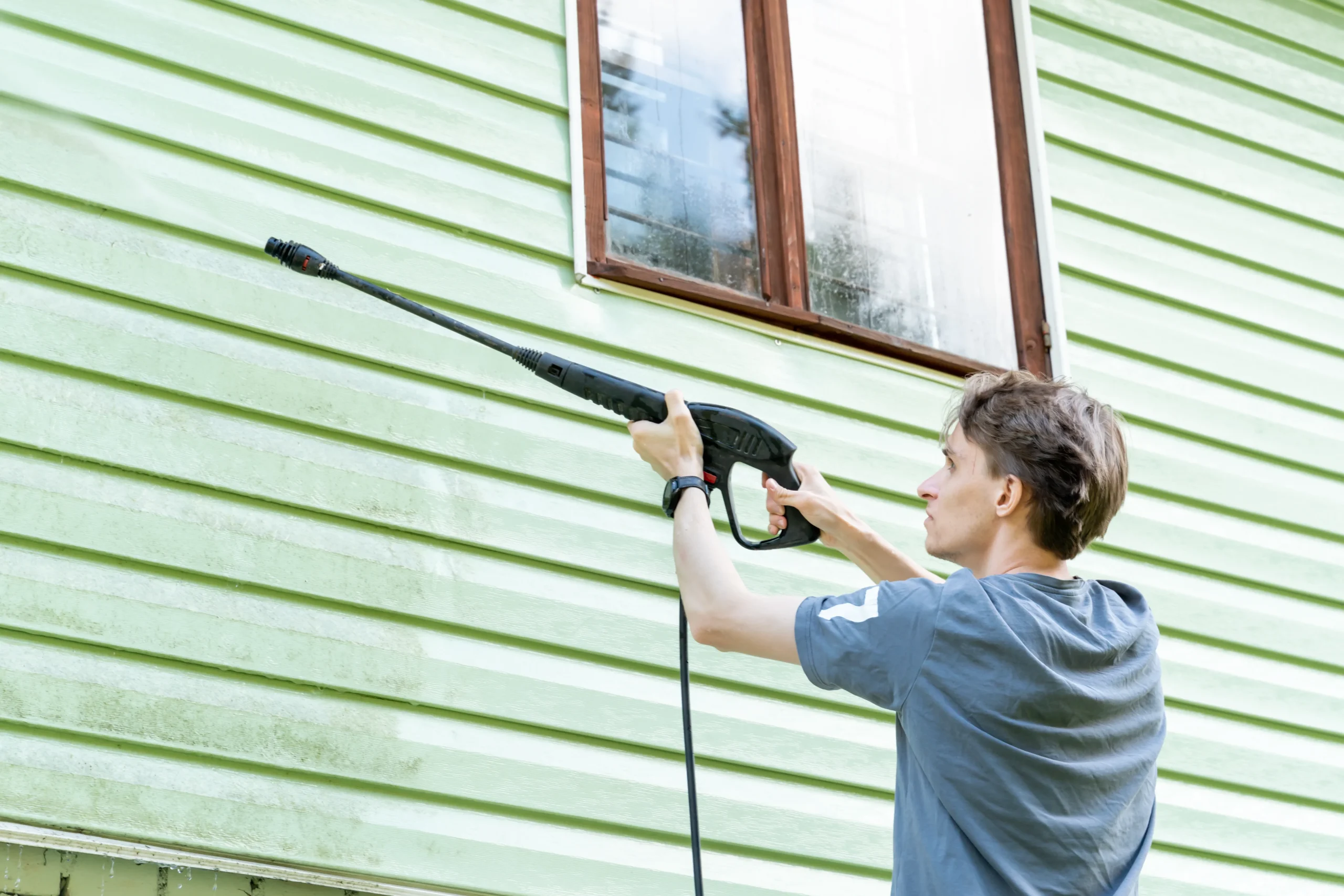 Nettoyage haute pression d'un bardage extérieur en préparation du support avant peinture ou rénovation de façade. Un artisan projette de l'eau sur les lames vertes à côté d'une fenêtre.